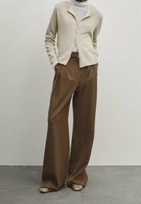 Cardigan beige con bottoni sopra un dolcevita grigio, abbinato a pantaloni marroni a gamba larga e scarpe beige su un pavimento grigio testurizzato.