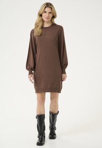 Abito lungo a maniche lunghe in maglia marrone con scollatura rotonda e polsini a coste, abbinato a stivali neri lucidi fino al ginocchio con fibbie.
