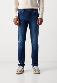 Dunkelblaue Denim-Jeans mit schmaler Passform, fünf Taschen, kupferfarbener Hardware und leichten Verwaschungen. Kombiniert mit grauen und beige Sneakers.