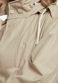 Beżowa kurtka z wysokim kołnierzem, zatrzaskami i regulowanym ściągaczem. Gładka tekstura i lekko luźny krój. Widoczne detale przeszyć.