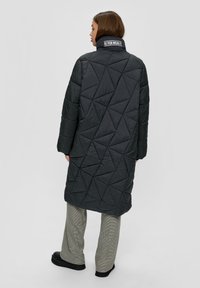Mujer vestida con un largo abrigo negro acolchado con un collar alto que presenta texto, pantalones de cuadros y zapatos negros, de pie de espaldas.