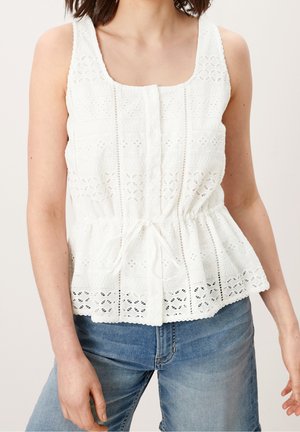 Vrouw draagt een mouwloze witte kanten blouse met knopen en taillekoord, gecombineerd met een blauwe spijkerbroek.