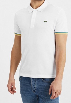 Homme portant un polo blanc à manches courtes avec des poignets rayés colorés et un petit logo vert sur la poitrine, assorti à un jean bleu.