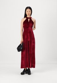 Burgundy bársonyos, nyakpántos overall kulcslyuk részlettel, széles lábszárral és illeszkedő derékkal. Fekete táskával és vastag talpú cipőkkel párosítva.