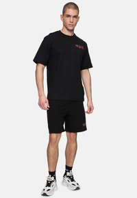 Sort bomulds t-shirt og shorts sæt med et lille rødt logo. Båret med hvide og sorte sportssko samt mønstrede sorte sokker.