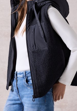 Person iført en sort tekstureret vest med frontlynlås og lomme over en hvid langærmet skjorte og lyseblå jeans.