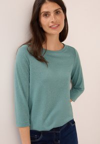 Jonge vrouw met lang donker haar en sproeten, gekleed in een teal lange mouwen top en donkere jeans, staand tegen een licht beige muur.