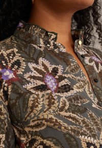 Camicia a fantasia floreale con collo. Presenta tonalità verde, viola e marrone. Realizzata in tessuto morbido con una finitura testurizzata e bottoni.