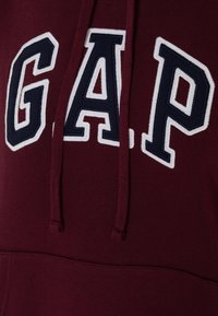 Sudadera color burdeos con el logo de "GAP" bordado en blanco y azul marino. Cuerdas de ajuste y bolsillo frontal visibles. Textura de tela suave evidente.