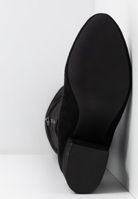 Stivale in suede nero con suola in gomma texturizzata e tacco basso, posizionato in verticale su uno sfondo bianco.