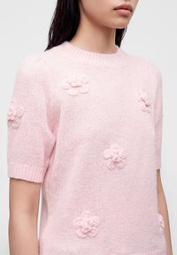 Γυναίκα που φοράει ανοιχτό ροζ κοντομάνικο πουλόβερ με ανάγλυφα 3D σχέδια λουλουδιών μπροστά και στα μανίκια, σε απλό φόντο.