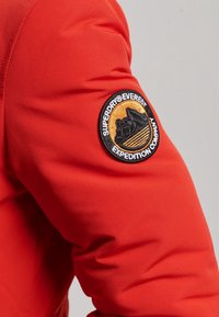 Czerwona rękaw kurtki z okrągłą naszywką "Superdry Everest Expedition Company" przedstawiającą sylwetkę góry i grafikę słońca.