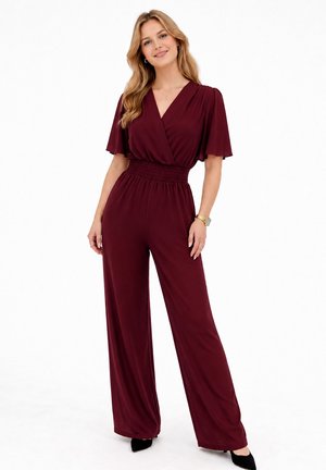 Kvinde med langt blond hår iført en burgundy jumpsuit med vidde ben, korte ærmer og elastisk talje, stående og smilende.