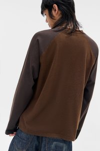 Νεαρό άτομο με μεσαίου μήκους σκούρα μαλλιά, φορώντας μακρυμάνικη καφέ και σκούρα γκρι μπλούζα και σκούρο μπλε τζιν, γυρισμένο προς τα πίσω.