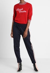 Roter Glitzerpullover mit "Soul Train" in Pink, kombiniert mit marineblauen Jogginghosen mit seitlichen Streifen und hochhackigen, lackierten Stiefeln.