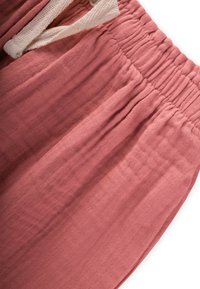 Kokvilnas bikses mīkstā rozā tonī ar teksturētu apdari. Elastīga jostasvieta ar baltu auklu. Iezīmē maigas krokas jostasvietā.