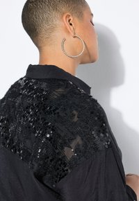 Maglietta nera con spalle decorate con paillettes, tessuto testurizzato e colletto. Gli orecchini sono grandi cerchi di argento.