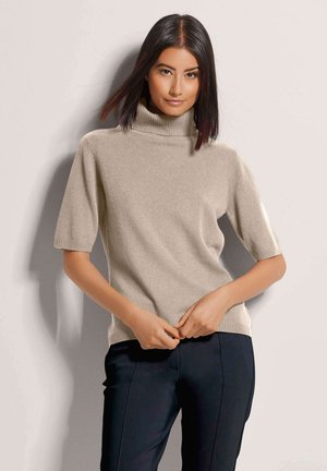 Vrouw met stijl donker haar, gekleed in een beige coltrui met korte mouwen en zwarte broek, staand tegen een lichtgekleurde muur, handen gevouwen.