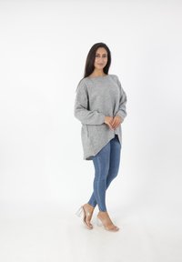 Lichtgrijze oversized trui met lange mouwen, een ronde hals en zijsplitsingen, gecombineerd met strakke blauwe jeans en doorzichtige blokkaneelsandalen.
