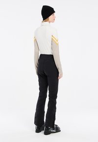 Langarm-Sportoberteil in Creme und Beige mit gelben Chevron-Streifen; schwarze Skihosen mit Reißverschlüssen an den Säumen; schwarze Mütze.