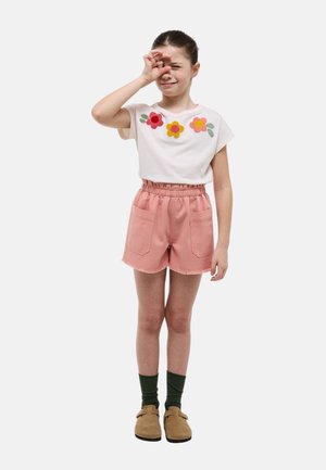Fille en chemise blanche à fleurs et short rose, fronçant les sourcils et se couvrant un œil avec la main, debout jambes droites.