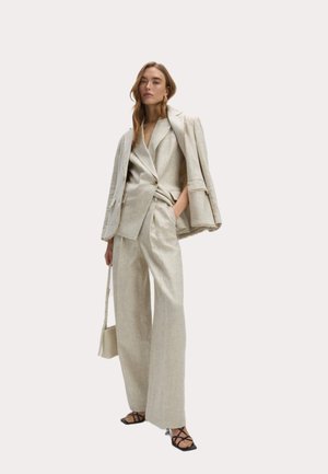 Traje de lino beige con un blazer entallado y pantalones de pierna ancha, que cuentan con detalles de botones y un cinturón en la cintura. Los accesorios incluyen un bolso pequeño.