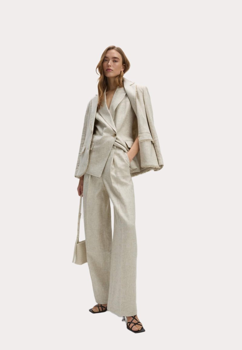 Completo in lino beige con giacca sartoriale e pantaloni a gamba larga, caratterizzati da dettagli con bottoni e cintura in vita. Gli accessori includono una piccola borsa.