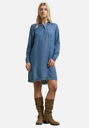 Woman with long blonde hair wears a loose blue denim dress with frayed hem and brown knee-high boots, standing against a plain background.