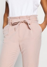 Ljust rosa byxor med bälte i midjan, prydda med randigt mönster, sidfickor och en slät yta. Stylat med en vit topp.