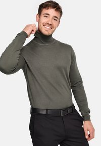 Dunkelgrüner Rollkragenpullover mit geripptem Muster, engen langen Ärmeln und hohem Kragen, kombiniert mit schwarzen Hosen und einem schwarzen Gürtel.