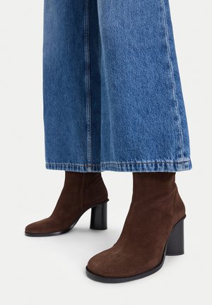 Stivaletti alla caviglia in camoscio marrone con tacchi a blocco indossati con jeans di jeans a gamba larga blu su uno sfondo bianco.