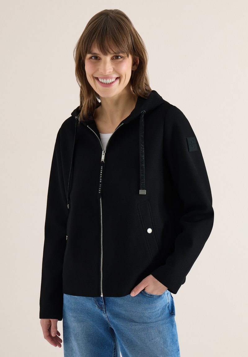 Femme souriante portant un sweat à capuche noir zippé avec cordons et un jean bleu, debout devant un fond clair uni.
