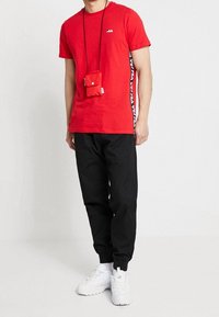 Camiseta roja de manga corta con una franja lateral negra que presenta un logo blanco; combinada con pantalones negros ajustados y zapatillas blancas; incluye un pequeño pouch rojo.