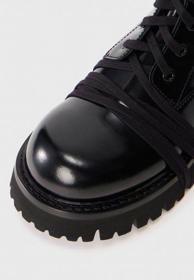 Bottine en cuir noir avec une finition brillante, bout rond, lacets en tissu et semelle en caoutchouc robuste avec un motif de crampons prononcé.
