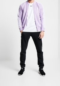 Ljuslila zip-up-jacka med vita ränder, tillsammans med en vit t-shirt och svarta träningsoverallsbyxor med vita strävfärgade detaljer. Svarta sneakers.