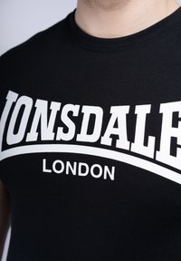 Zwarte katoenen t-shirt met gedurfde witte tekst die "LONSDALE LONDON" in een gebogen ontwerp over de voorkant leest.