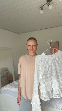 Giovane donna che tiene una camicetta bianca di pizzo su una gruccia in una luminosa camera da letto con letto e specchio sullo sfondo.