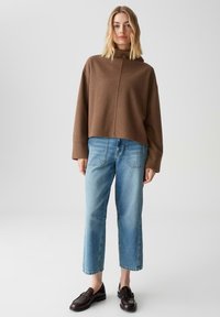 Maglione corto marrone con collo alto, abbinato a jeans a gamba dritta di colore blu chiaro. Il modello indossa mocassini scuri e ha i capelli acconciati in onde morbide.