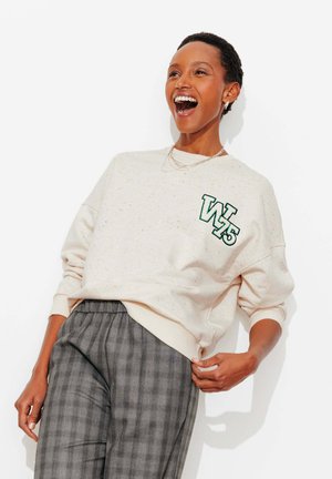 Femme riant portant un sweat-shirt clair à motifs tachetés avec le logo "W 75" et un pantalon à carreaux gris, posant devant un fond blanc.