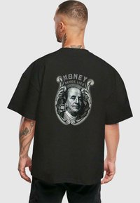 Fekete, nagyméretű póló, amelyen Benjamin Franklin grafikája található, fölötte a "A PÉNZ SOHA NEM HAZUDIK" felirattal, egy díszítő kerettel.