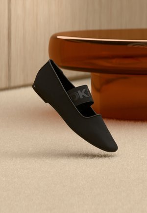 Black pointed-toe flat shoe with an elastic strap across the top, positioned on a beige carpet near a brown rounded table base.