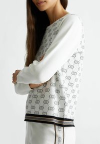 Maglione bianco con motivo circolare grigio, polsini e orlo a costine, caratterizzato da una banda accentuata nera e dorata. Tessuto liscio, vestibilità comoda.