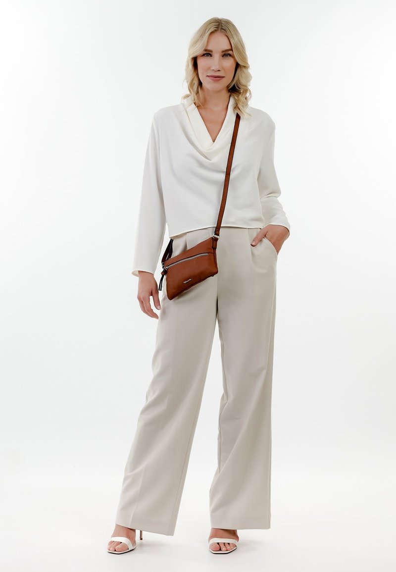 Femme blonde portant un haut blanc à manches longues, un pantalon beige à jambes larges, des talons blancs et un sac bandoulière marron, debout devant un fond blanc.