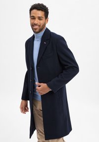 Marineblauer Wollüberrock, einreihiges Design mit vier Knöpfen, mit Reverskragen und einer Brusttasche. Kombiniert mit einem hellblauen Rollkragenpullover.