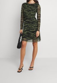 Mustekka ja vihreä tiikeriraidoitusmekko pitkillä läpinäkyvillä hihoilla, pyöreällä kaula-aukolla, drapeeratuilla yksityiskohdilla ja levenevällä hameella; yhdistetty mustaan käsilaukkuun.