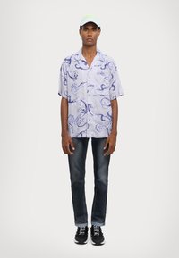 Hombre vestido con una camisa de botones de manga corta de color púrpura claro con estampado, jeans oscuros, zapatillas negras y una gorra blanca con letras en neón verde.