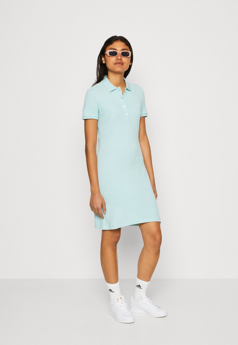 Donna che indossa un vestito polo blu chiaro, calze bianche Adidas, sneaker bianche e occhiali da sole rettangolari, in piedi su uno sfondo neutro.