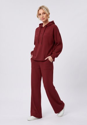Femme debout portant un sweat à capuche bordeaux et un pantalon large assorti avec des baskets blanches sur un fond uni.