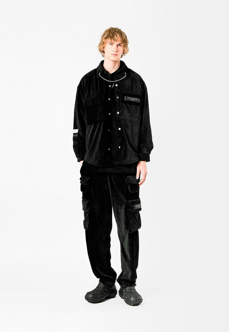 Chaqueta de pana negra con bolsillos con botón y cremallera, combinada con pantalones cargo que presentan múltiples bolsillos. El conjunto incluye calzado texturizado.