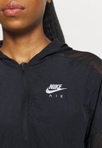 Svart lättviktsjacka med huva, tillverkad av strukturerad tyg, med dragkedja och en framträdande vit Nike Air-logotyp på bröstet.